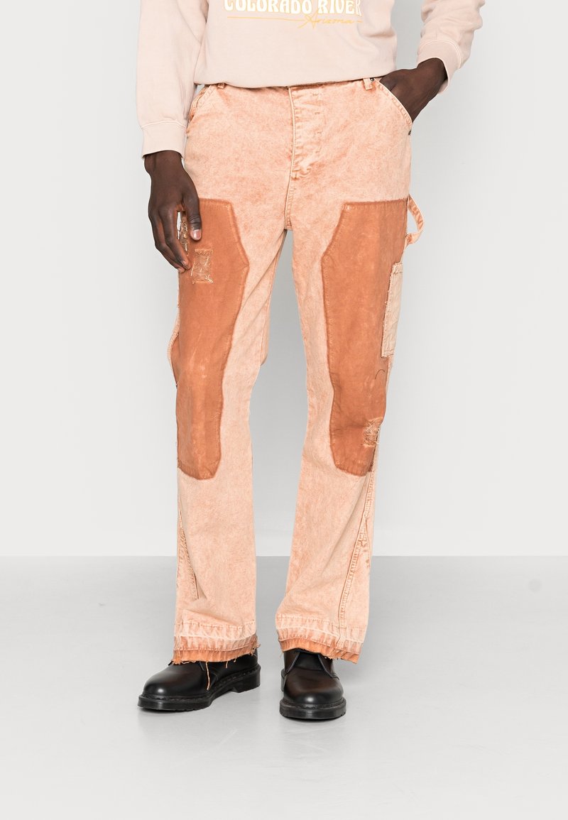Persona que lleva pantalones de un naranja claro con parches de naranja más oscuro en los muslos, combinados con zapatos negros y una sudadera beige con texto.