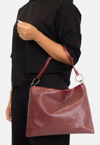 Bolso de mano texturizado en rojo con tachuelas metálicas, que cuenta con un cierre de cremallera y una sola correa de hombro sujeta con herrajes plateados.