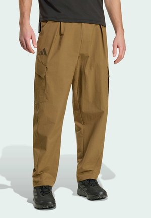 Uomo che indossa pantaloni cargo marroni con tasche laterali e scarpe sportive nere, in piedi contro uno sfondo chiaro e uniforme.