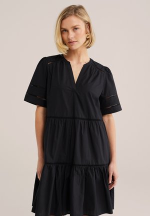 Lühikeste blondide juustega naine, kes kannab musta mitmetasandilist kleiti lühikeste varrukatega ja dekoratiivsete lõike detailidega, seisab ühevärvilise tausta ees.