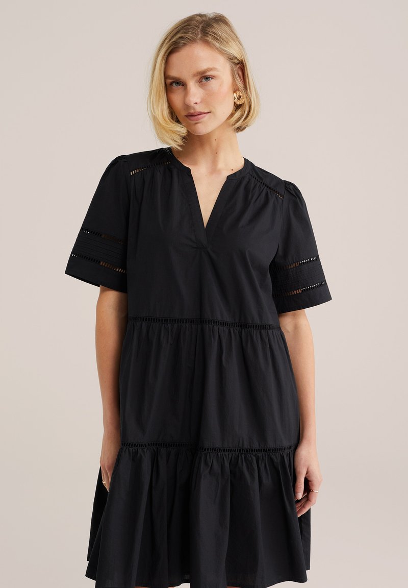 Femme aux cheveux blonds courts portant une robe noire à volants avec manches courtes et détails décoratifs ajourés, posant devant un fond uni.
