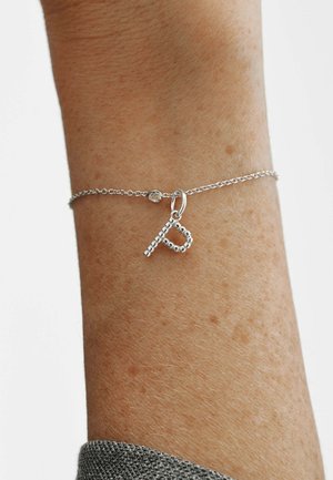 Pulsera delicada de cadena de plata con un colgante de cuentas en forma de letra "B" sobre una muñeca, sobre un fondo liso.