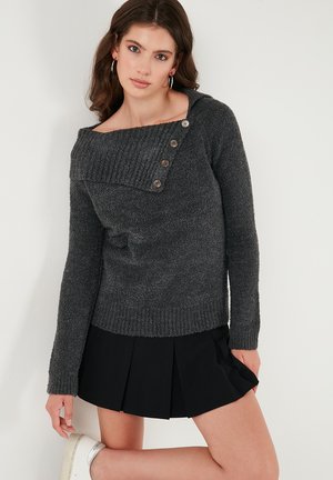 Grijze trui met een brede off-shoulder halslijn, geribbelde textuur en een drie-knopen detail op de schouder, gecombineerd met een zwarte, geplooide rok.