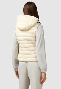 Lichtbeige puffer vest met een capuchon, gewatteerd ontwerp en elastische zoom. Gepaarde met een lichtgrijze langslovende kledingstuk en bijpassende broek.