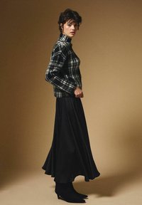 Kobieta w czarno-białym swetrze w kratkę z golfem, długiej czarnej spódnicy i czarnych botkach na obcasie stojąca na jednolitym beżowym tle.