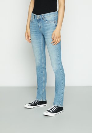 Person trägt hellblaue Slim-Fit-Jeans, schwarze Low-Top-Sneaker und ein schwarzes ärmelloses Oberteil und steht vor einem schlichten Hintergrund.
