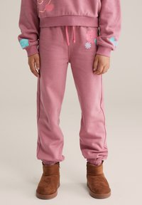 Lyserøde joggingbukser af blødt stof med en løs pasform, prydet med blomster- og smilende ansigtsmotiver og elastiske manchetter. Bruges med brune ankelstøvler.