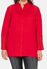 Blouse rouge avec col mao, patte de boutonnage à l'avant et plis nervurés. Manches transparentes à pois qui ajoutent de la texture au corps uni.