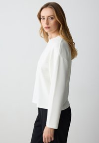 Maglia bianca a maniche lunghe con scollo rotondo, caratterizzata da decorazioni in strass sparse e una vestibilità rilassata, abbinata a pantaloni neri.
