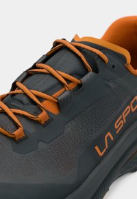 Tmavě šedá běžecká bota s oranžovými akcenty, má texturovaný svršek, výrazné tkaničky a detail loga na boku. Lehký design.