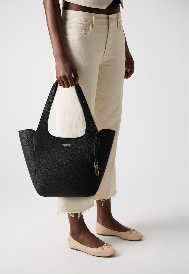 Zwarte gestructureerde tote bag met dubbele handvaten, gouden logo-detail en kleine slotaccent. Model draagt een crème cropped broek en beige flats.