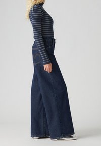 Breda ben mörka denimjeans som bärs med en långärmad marinblå randig topp. Jeanssen har en klassisk design med fem fickor och synliga sömmar.