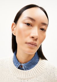 Nahaufnahme einer Person, die einen cremefarbenen Strickpullover und ein blaues Jeanshemd trägt, wobei ein strukturiertes Strickmuster und die geschichteten Kragen zur Geltung kommen.