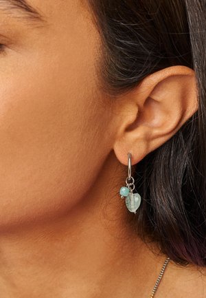Gros plan sur une oreille avec une boucle d'oreille argentée en forme de créole ornée de deux pierres précieuses bleu clair pendantes.