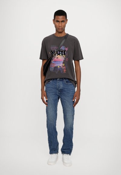 Grafisches T-Shirt in Dunkelgrau mit buntem Druck, der Landschaften und abstrakte Designs zeigt, kombiniert mit blauen Jeans und weißen Sneakers.
