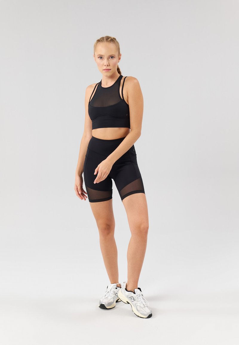 Svart treningssett med en tettsittende sports-bh med meshpaneler og høye shorts med mesh-detaljer, kombinert med sølvfargede joggesko.