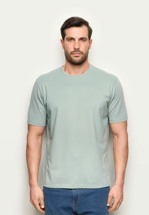 Uomo con capelli corti e scuri e barba, che indossa una maglietta a maniche corte grigio chiaro semplice e jeans blu, in piedi davanti a uno sfondo bianco.