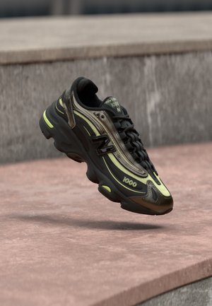 Černo-žlutá teniska New Balance 1000 zavěšená nad růžovým kamenným povrchem s pozadím šedé kamenné zdi.