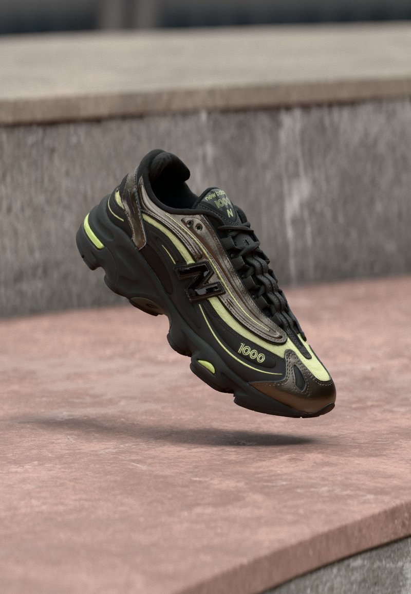 Μαύρο και κίτρινο αθλητικό παπούτσι New Balance 1000, κρεμασμένο πάνω από επιφάνεια από ροζ πέτρα, με φόντο γκρι πέτρινο τοίχο.