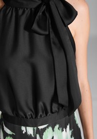 Top en satin noir avec un grand nœud au cou, taille froncée et texture lisse, associé à une partie inférieure à motifs en vert et noir.