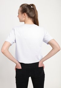 Hvid bomulds T-shirt med korte ærmer, rund hals, afslappet pasform, båret med sorte jeans med baglommer. Set fra ryggen.