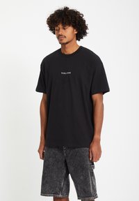 Svart bomull t-skjorte med korte ermer og rund hals, med en hvit "VOLCOM" logo på brystet. Kombinert med grå denimbukser.