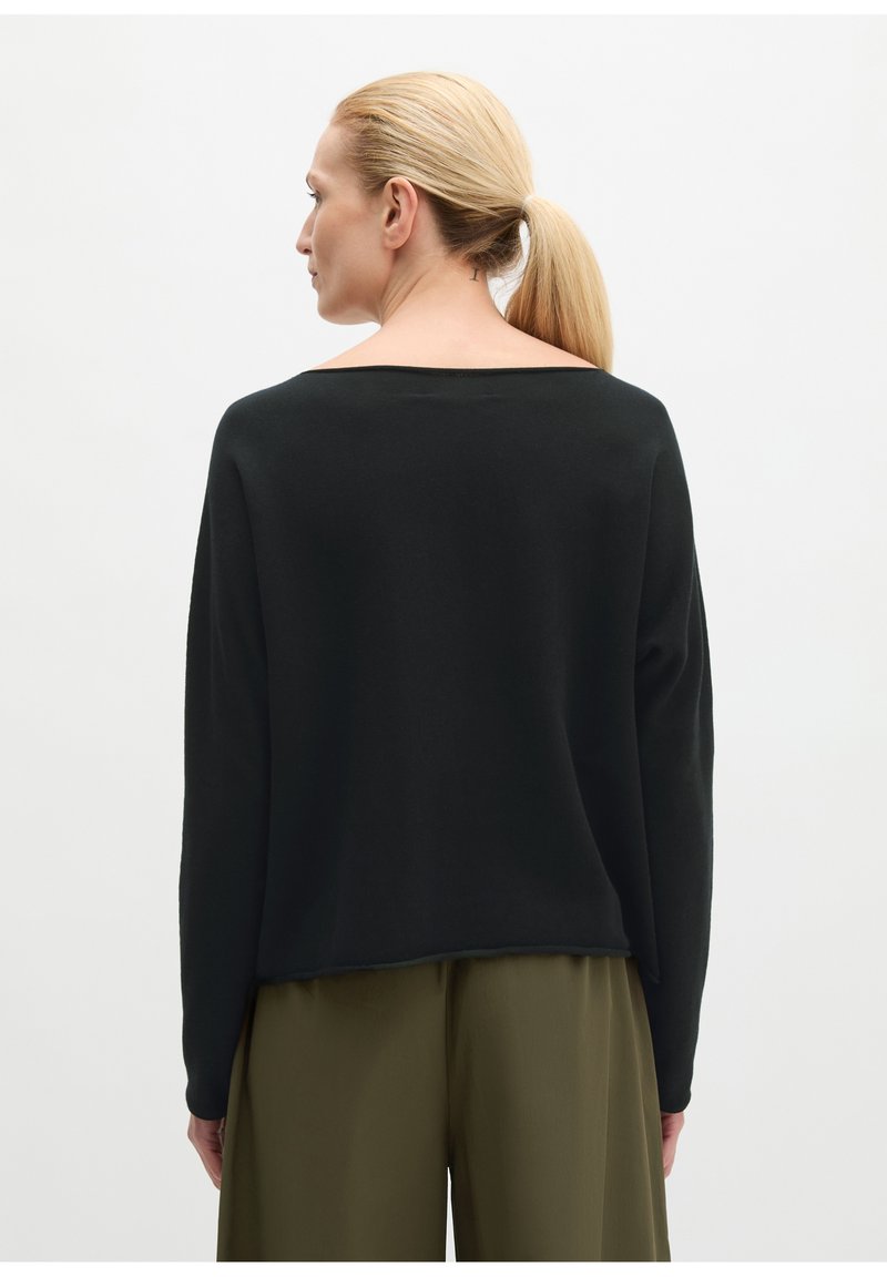Femme avec queue de cheval blonde portant un haut noir ample à manches longues et un pantalon large vert olive, vue de dos sur un fond blanc uni.