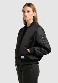 Bomber jacket nera realizzata in materiale lucido. Caratterizzata da una tasca con zip sulla manica, polsini a coste e un'etichetta con logo sull'orlo inferiore.