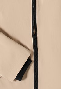 Manche de veste beige avec revers intérieur noir à côté d'une fermeture éclair noire partiellement ouverte sur un tissu beige.