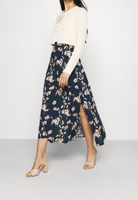 Kremowy, prążkowany krótki kardigan z detalem w postaci wiązania, zestawiony z granatową, kwiecistą spódnicą maxi z bocznym rozcięciem i różowymi wzorami kwiatów.