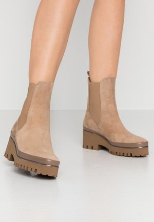 Beige ruskind ankelstøvler med elastiske sidedele, forhøjet chunky hæl og mønstret gummisål, med en afrundet tådesign.