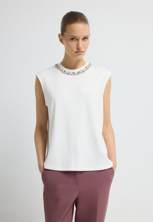 Femme avec les cheveux lissés en arrière portant un haut sans manches blanc avec un col en pierres précieuses et un pantalon mauve ajusté, les mains dans les poches.