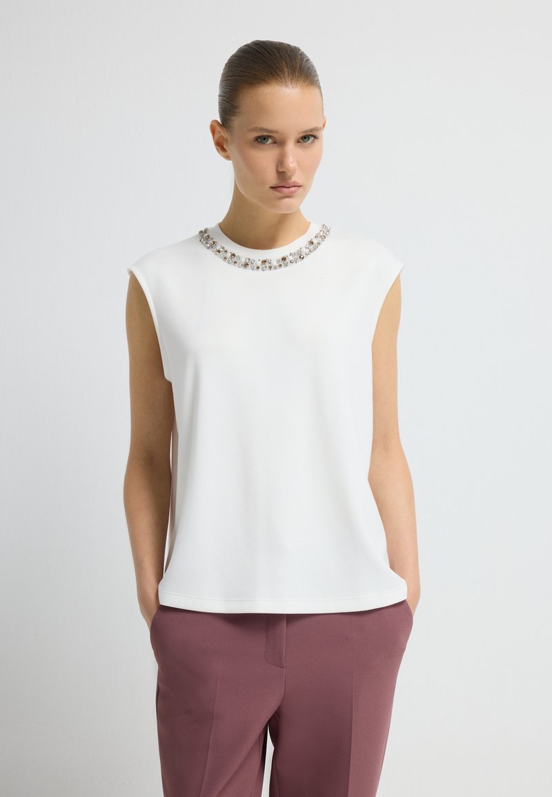 Frau mit zurückgegelten Haaren, die ein ärmelloses weißes Oberteil mit einem mit Edelsteinen besetzten Ausschnitt und mauve-farbene, taillierte Hose trägt, mit den Händen in den Taschen.