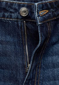 Jean en denim avec fermeture éclair métallique visible à moitié ouverte et bouton gravé du nom de la marque, avec détails de couture orange.