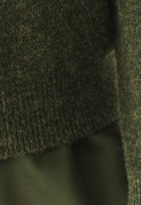 Svetr z příze v tmavě olivově zelené barvě, s texturovaným, chlupatým povrchem. Spodní lem je ribbovaný; doplněný hladkým, sladěným materiálem pod ním.