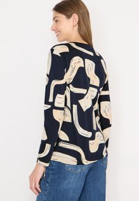 Nainen, jolla on yllään pitkähihaisen laivastonsininen paita, jossa on beigejä abstrakteja siveltimenvetoja, yhdistettynä sinisiin farkkuihin, seisoo valkoista taustaa vasten.