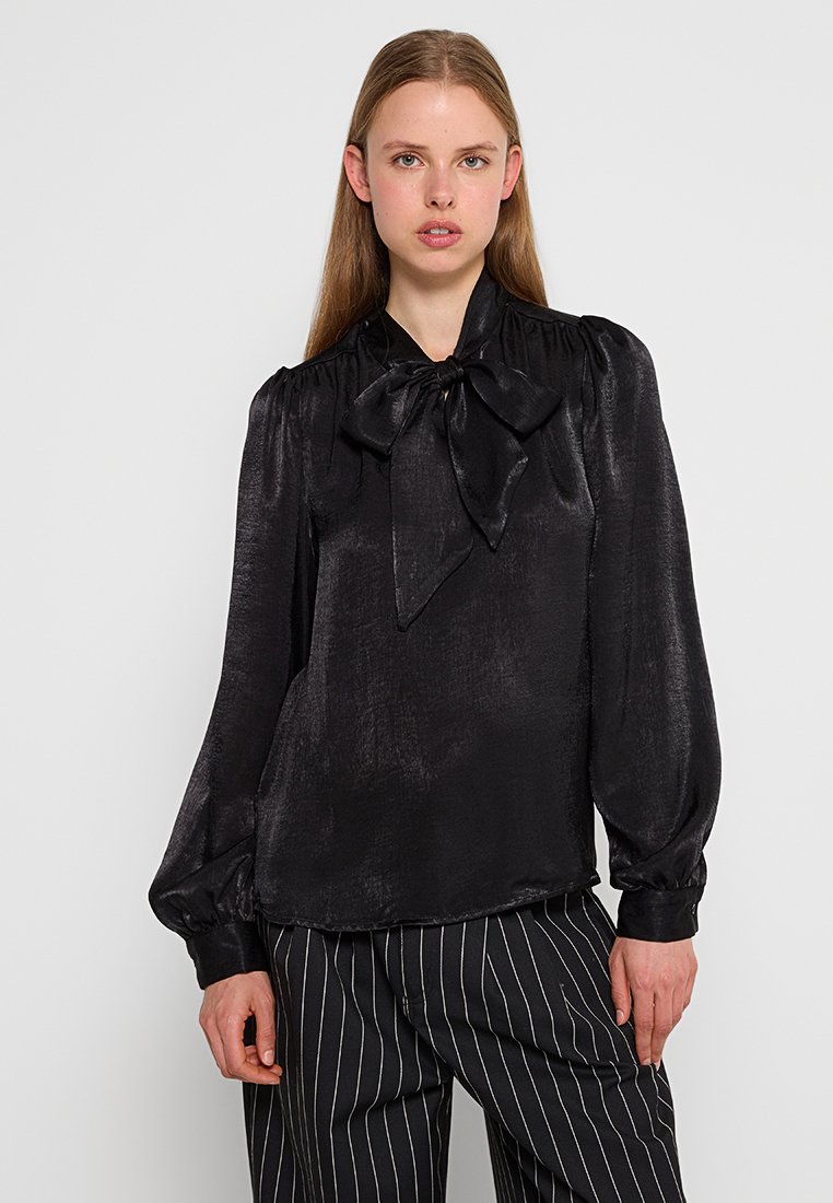 Blusa negra de satén con un cuello alto, gran lazo decorativo y mangas abullonadas. La tela tiene una textura sutilmente brillante.
