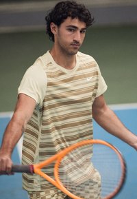 Mladý muž v béžovém pruhovaném tričku držící oranžovou tenisovou raketu na vnitřním modrém tenisovém kurtu, soustředěný na hru.