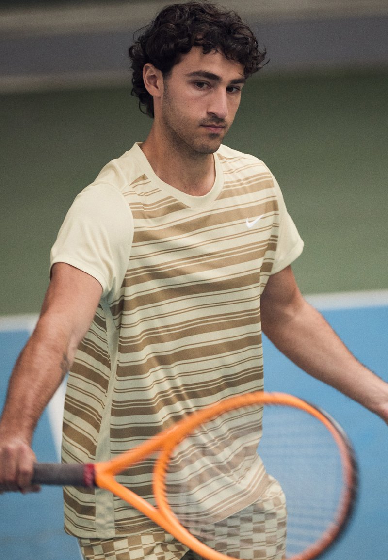Herre tennisantrekk i lysegul og brunstripet stoff. Har rund hals, korte ermer, og holder en oransje racket.