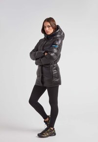 Czarna puchówka z kapturem, o błyszczącej fakturze, z troczkiem w talii i kieszeniami zapinanymi na zamek. Zestawiona z czarnymi legginsami i butami sportowymi.