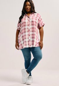 Camicia a quadri rosa chiaro e bianca con maniche corte, chiusura a bottoni e vestibilità rilassata, abbinata a jeans skinny blu e sneakers bianche.