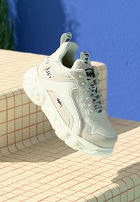 White and light gray sneaker featuring mesh and synthetic materials, chunky sole, lacing detail, and branding on the heel and tongue.