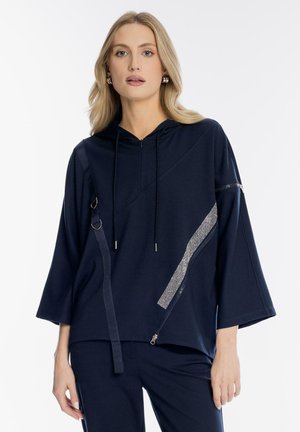 Blondynka w granatowej bluzie z szerokimi rękawami, suwakiem oraz dopasowanych granatowych spodniach, stojąca na białym tle.