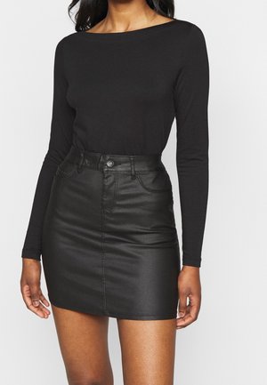 Vrouw die een nauwsluitende zwarte top met lange mouwen en een hoge taille zwarte leren minirok draagt, tegen een effen achtergrond.