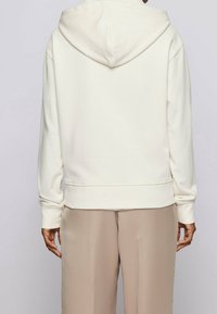 Sudadera blanca con una textura suave, que presenta una capucha con cordón, hombros caídos y puños de canalé. Llevada sobre pantalones beiges.