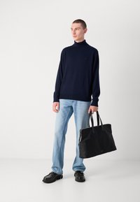 Marinefarbener Rollkragenpullover, hellblaue Jeans und schwarze Lederhandtasche. Die Tasche hat zwei Griffe und einen Reißverschluss. Die Schuhe sind schwarze, klobige Schuhe.