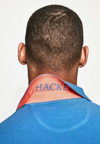 Modrá polo košile s kontrastním oranžovým vnitřním límcem, na němž je modrý text "HACKER". Viditelné textury a detaily šití na látce.