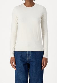 Långärmad topp i krämfärg, tillverkad av mjukt material med rund halsringning och slät yta, tillsammans med mörkblå denimjeans.