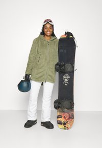 Grüne Fleece-Jacke mit Reißverschluss, weiße Schneehose, schwarzes Snowboard mit Grafikdesign und Bindungen; schwarzer Helm und Handschuhe in der Hand.