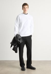 Sudadera blanca de cuello redondo, con logo negro, combinada con pantalones negros y una chaqueta de cuero colgada del brazo. Textura suave, diseño minimalista.